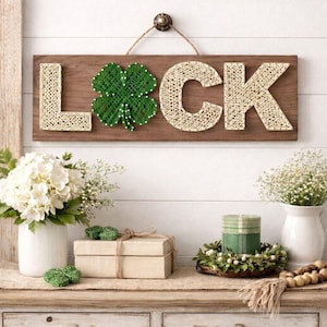 Może przedstawiać: Drewniany znak z napisem "LUCK" wykonanym techniką string art. Litery "L" i "C" są beżowe, "K" jest beżowe, a "U" to zielona czterolistna koniczyna. Znak wisi na białej ścianie. Przedmioty dekoracyjne znajdują się na drewnianym stole.
