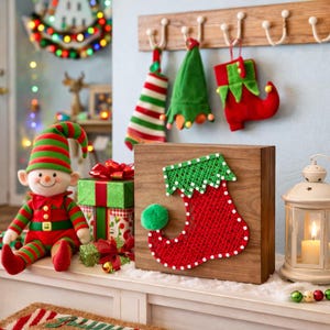 Puede incluir: Una exhibición navideña festiva con un muñeco de elfo, cajas de regalo y medias decorativas. Un bloque de madera muestra una media de arte de hilo. La paleta de colores incluye rojo, verde y blanco, creando un ambiente festivo alegre.