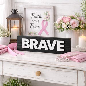 以下が含まれることがあります： 「BRAVE」の白い文字が書かれた黒い木製の看板が特徴的な装飾。 「Faith Over Fear」の文字とピンクのリボンが入った額入りのプリントが見えます。 ピンクの花、キャンドル、ランタンがシーンを完成させています。