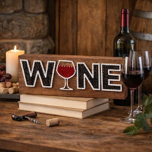May include: A wooden sign with the word "WINE" spelled out in black letters with white stitching. A wine glass graphic is in the center. Two wine glasses filled with red wine and a bottle of wine are in the background.