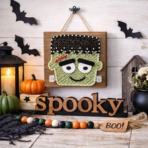 May include: A Halloween string art of Frankenstein's monster on a wooden board. The word "spooky" is displayed in wooden letters. Decorative bats, pumpkins, and a "BOO!" tag complete the festive scene.