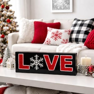 May include: A black wooden sign with the word "LOVE" in red letters and a white snowflake. The letters are outlined with a red glitter material. The sign is on a white table with a Christmas tree and decorative pillows in the background.