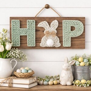Op de afbeelding: Houten bord met het woord "HOP" en een konijnontwerp. De letters "H" en "P" zijn lichtgroen en het konijn is wit met een bruine strik. Het bord hangt aan een witte muur, met paasdecoraties op de voorgrond.