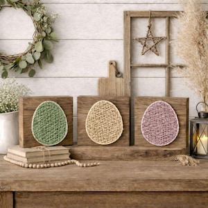 Puede incluir: Tres decoraciones de huevos de Pascua de madera. Cada huevo está hecho de cuerda verde, beige y rosa. Los huevos se exhiben sobre una mesa de madera con otras decoraciones rústicas, incluida una corona.