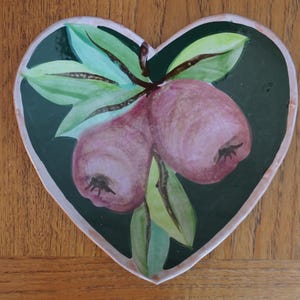 May include: Heart-shaped ceramic plate with a hand-painted design of two pinkish-red pears and green leaves on a dark green background. The plate has a white border and is set against a wooden surface.