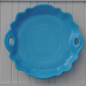 May include: A bright turquoise round serving tray with a scalloped edge and two integrated side handles. The center of the tray features a subtle embossed circular pattern, and the overall design has a smooth, glossy finish throughout the piece.