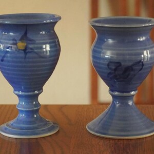 May include: Two blue ceramic goblets with a flared rim and a pedestal base. Each goblet features a horizontal ribbed texture along the body and a hand-painted dark blue floral motif. The glaze has a glossy finish with subtle variations in color.