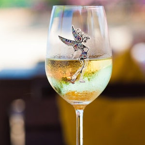 May include: A wine glass filled with a light-colored liquid, possibly white wine, with a decorative fairy charm attached. The fairy charm has colorful accents and is positioned on the side of the glass.