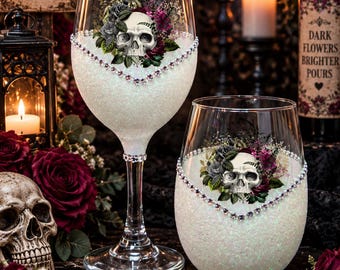 May include: Two clear glass wine vessels, one stemmed and one stemless, featuring a white glitter base, a rhinestone trim, and a central graphic of a human skull surrounded by dark red and black roses. The glass surfaces are smooth and transparent.