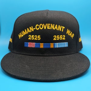 Könnte beinhalten: Eine schwarze Baseballkappe mit dem Text "HUMAN-COVENANT WAR" in gelber Stickerei. Die Zahlen "2525" und "2552" sind ebenfalls in Gelb gestickt, zusammen mit einer Reihe von fünf bunten Streifen.