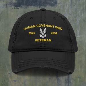 Könnte beinhalten: Eine schwarze Baseballkappe im Used-Look mit gelber Stickerei, die HUMAN-COVENANT WAR, 2525, 2552 und VETERAN zeigt. In der Mitte befindet sich ein weißes gesticktes Emblem eines geflügelten Adlers, der eine Weltkugel mit dem Schriftzug UNSC hält.