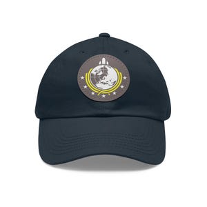 May include: A navy blue baseball cap featuring a circular patch on the front. The patch is grey with a white graphic of the Earth, a rocket, and a yellow curved line with five white stars arranged in a semi-circle along the bottom edge.