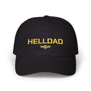 Puede incluir: Una gorra de béisbol negra con el texto HELLDAD bordado en amarillo centrado en el panel frontal. Debajo del texto hay un pequeño emblema bordado en amarillo que consiste en un icono alado estilizado con una forma central de casco.