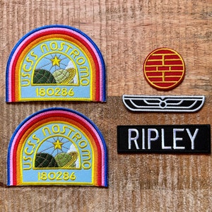 May include: A selection of embroidered patches on a wooden surface. Two patches display a rainbow arch design with the text "USCSS NOSTROMO" and the number "180286". Other patches include a red and gold circular design, a black and white emblem, and a black patch with the name "RIPLEY".