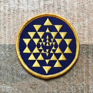 Op de afbeelding: Een marineblauwe geborduurde patch met een gouden geometrisch ontwerp. Het ontwerp is een herhalend patroon van driehoeken die een piramidevorm vormen.