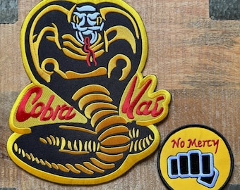 Puede incluir: Dos parches bordados, uno con una cobra amarilla y las palabras "Cobra Kai" en rojo y amarillo, y el otro con un puño amarillo y las palabras "No Mercy" en negro.
