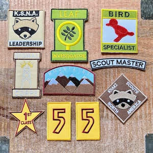 Puede incluir: Una colección de diez parches bordados con varios diseños y textos. Los parches son de varios colores, incluyendo verde, amarillo, marrón y rojo. Los parches presentan texto como "K.S.N.A. Leadership", "Leaf Investigator", "Bird Specialist", "Signal Corps", "Mountains", "Scout Master", "Khaki Scouts North America", "1st Class", y "55".