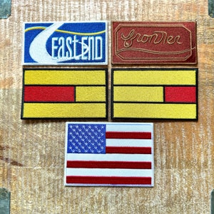 Könnte beinhalten: Fünf gestickte Patches, darunter ein blauer Patch mit weißem Text, der "East End" sagt, ein roter Patch mit weißem Text, der "Growler" sagt, zwei gelbe Patches mit roten Streifen und ein rot-weiß-blauer amerikanischer Flaggen-Patch.