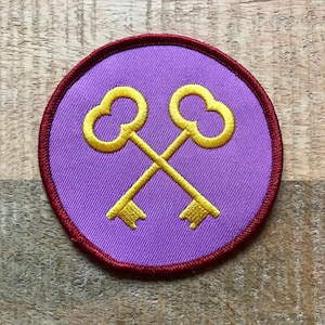 May include: A purple embroidered patch with a red border, featuring two gold crossed keys.