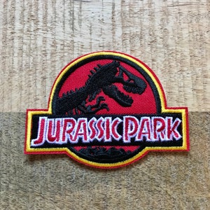 Puede incluir: Un parche bordado rojo, amarillo y negro con el logotipo de Jurassic Park. El logotipo presenta una silueta negra de un dinosaurio con las palabras "Jurassic Park" en letras blancas con un contorno rojo.