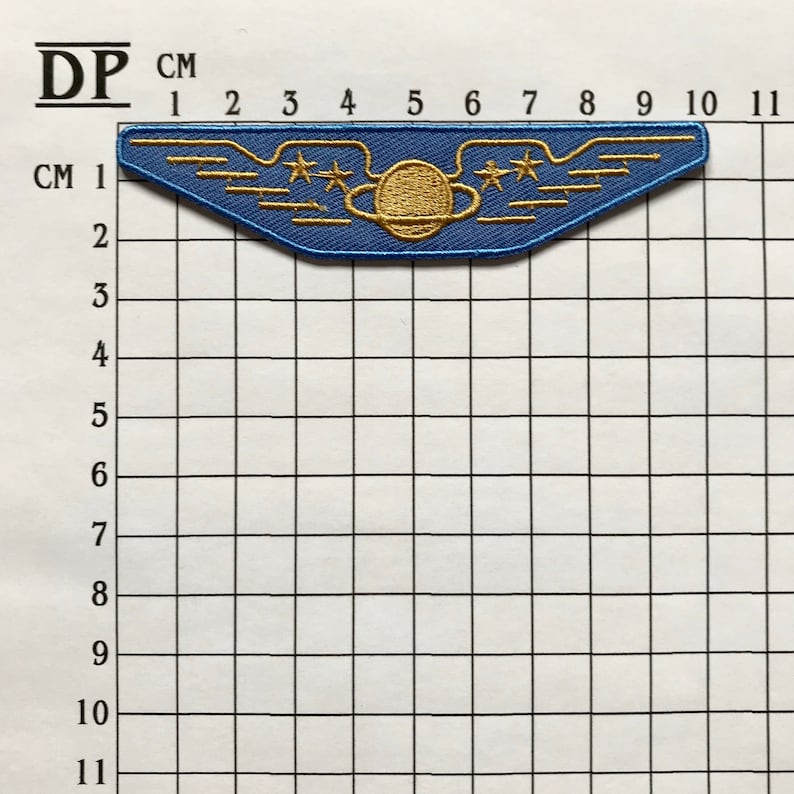 May include: Blue embroidered patch with gold wings, a planet, and three stars. The patch is approximately 5 cm wide and 2 cm tall.
