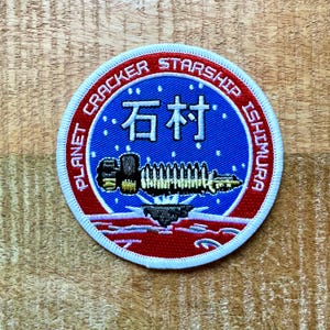 Op de afbeelding: Een blauw en rood geborduurde patch met witte tekst die "Planet Cracker Starship Ishimaru" en "石村" luidt. De patch toont een gestileerd ruimteschip met een wit en gouden ontwerp.