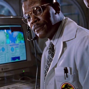 Op de afbeelding: Een man met een bril en een headset, gekleed in een witte laboratoriumjas met een "Jurassic Park" logo-patch. Hij kijkt naar een computerscherm met een kaart. Er zit een pen in de jaszak.