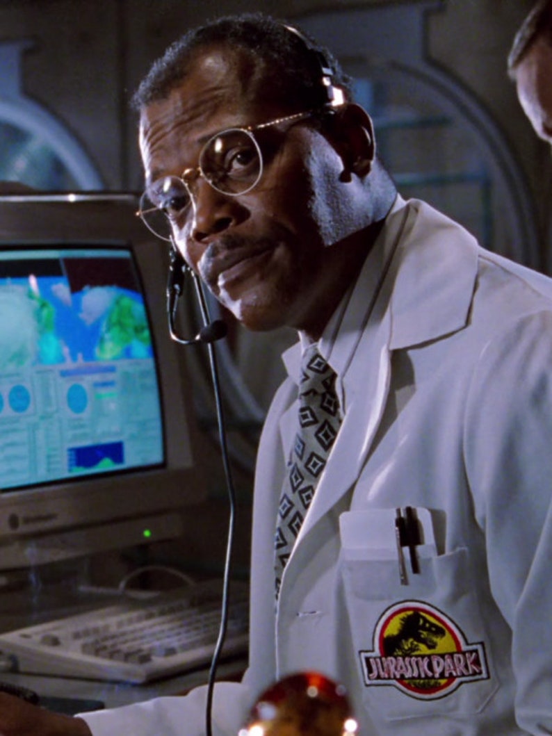Op de afbeelding: Een man met een bril en een headset, gekleed in een witte laboratoriumjas met een "Jurassic Park" logo-patch. Hij kijkt naar een computerscherm met een kaart. Er zit een pen in de jaszak.
