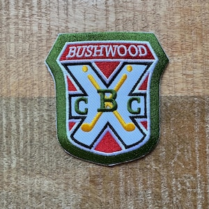 May include: Embroidered patch with a shield design. The patch features the word "BUSHWOOD" in white on a red banner. The shield has a green border, white background, and crossed golf clubs. The letters "CBC" are also visible.
