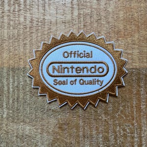 Könnte beinhalten: Ein gestickter Aufnäher in Gold und Weiß mit dem Text "Official Nintendo Seal of Quality".