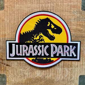 Op de afbeelding: Geborduurde patch met het Jurassic Park-logo. Het ontwerp toont een zwart dinosaurus skelet op een gele achtergrond, een rood-witte rand en de woorden "JURASSIC PARK" in wit op een zwarte rechthoek. Retro film thema.