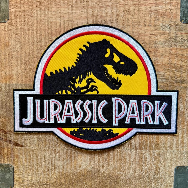 Op de afbeelding: Geborduurde patch met het Jurassic Park-logo. Het ontwerp toont een zwart dinosaurus skelet op een gele achtergrond, een rood-witte rand en de woorden "JURASSIC PARK" in wit op een zwarte rechthoek. Retro film thema.