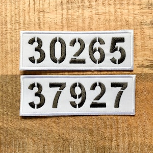 Puede incluir: Dos parches rectangulares blancos con números bordados en negro. El parche superior tiene los números "30265" y el parche inferior tiene los números "37927".
