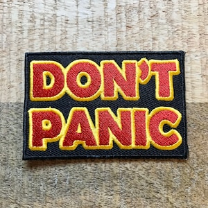 Könnte beinhalten: Ein schwarzer Stoff-Patch mit gelber Naht um die Ränder. Der Patch trägt die Worte "DON'T PANIC" in roten Buchstaben mit gelber Umrandung.