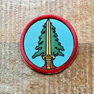Może przedstawiać: Okrągła naszywka haftowana z czerwonym obramowaniem, przedstawiająca zielone drzewo sosnowe z złotym mieczem na niebieskim tle.