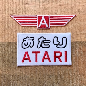 Peut inclure: Deux patchs brodés, l'un rouge avec un 'A' blanc et des ailes, l'autre blanc avec des caractères japonais noirs et le texte 'ATARI' rouge.
