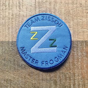 Puede incluir: Un parche circular azul con costuras blancas. El parche tiene el texto "Team Zissou Master Frogman" alrededor del borde y una gran letra blanca "Z" en el centro. La letra "Z" tiene una "Z" amarilla y una "Z" verde que se superponen.