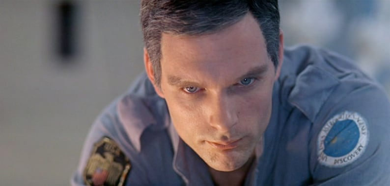 May include: A close-up of a man's face with a serious expression. He is wearing a blue shirt with a patch that says "United States Space Agency, Mission Discovery".