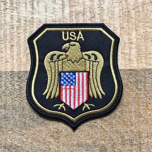 May include: A black and gold embroidered patch with a shield shape. The shield features a bald eagle with outstretched wings holding an American flag in its talons. The text "USA" is above the eagle.
