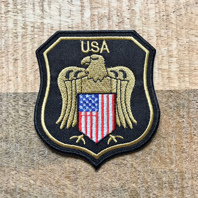 May include: A black and gold embroidered patch with a shield shape. The shield features a bald eagle with outstretched wings holding an American flag in its talons. The text "USA" is above the eagle.
