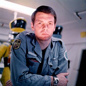 May include: A man wearing a blue jumpsuit with a patch that says "POOLE'S" and "Mariner" on it. He is standing in front of a yellow and black robot.
