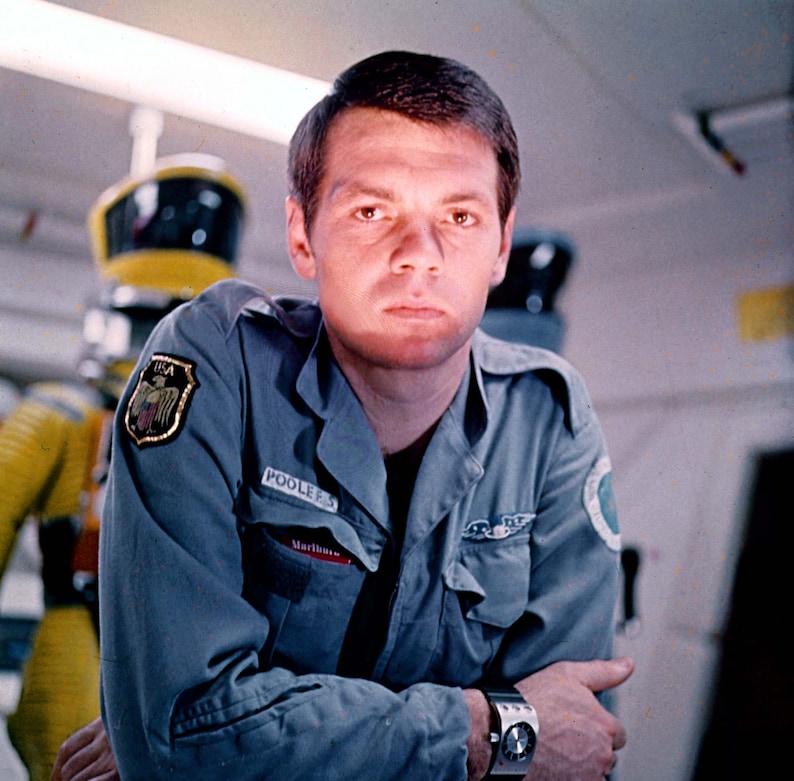 May include: A man wearing a blue jumpsuit with a patch that says "POOLE'S" and "Mariner" on it. He is standing in front of a yellow and black robot.