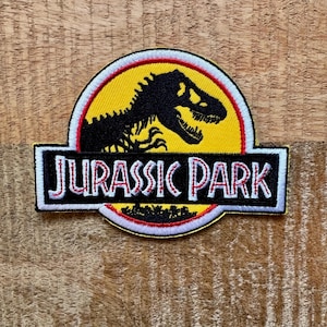 Op de afbeelding: Geborduurde patch met het "Jurassic Park" logo. Het ontwerp bevat een zwart silhouet van een Tyrannosaurus Rex op een gele achtergrond, met de woorden "JURASSIC PARK" in witte letters op een zwarte rechthoek, omlijnd in rood en wit.