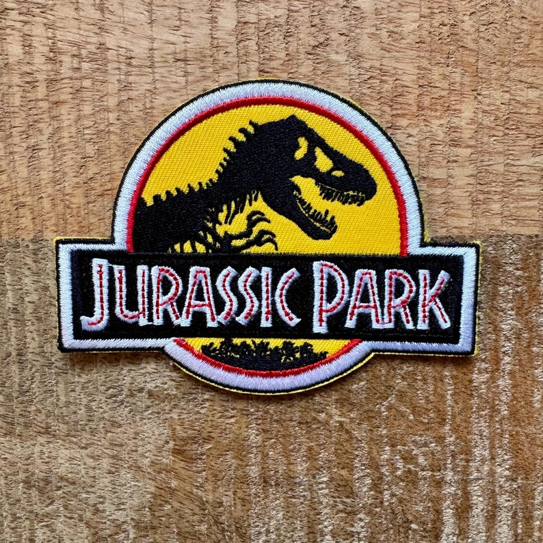 Op de afbeelding: Geborduurde patch met het "Jurassic Park" logo. Het ontwerp bevat een zwart silhouet van een Tyrannosaurus Rex op een gele achtergrond, met de woorden "JURASSIC PARK" in witte letters op een zwarte rechthoek, omlijnd in rood en wit.