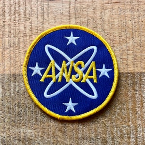Puede incluir: Un parche bordado azul y amarillo con el texto "AWSA" en letras amarillas rodeado de un símbolo atómico blanco y cuatro estrellas blancas.