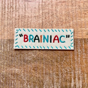 May include: A white fabric patch with blue stitching around the edges. The patch has the text "BRAINIAC" embroidered in red, orange, black, and blue thread.