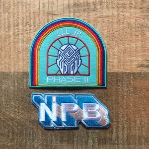 Peut inclure: Deux patchs brodés. Le premier patch est bleu avec un arc-en-ciel, un cercle blanc avec un motif en forme de labyrinthe à l'intérieur et le texte "ULP PHASE III". Le deuxième patch est bleu avec le texte "NPB" en blanc.