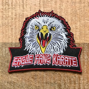 May include: An embroidered patch with a fierce eagle head with red, white, and yellow details. The eagle's beak is open and its eyes are wide. The patch has a black background with a red banner that reads "Eagle Fang Karate".
