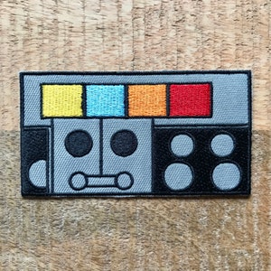 Może przedstawiać: Szary haftowany znaczek z twarzą robota. Robot ma dwa czarne oczy, czarne usta i cztery czarne guziki. Górna część robota ma cztery kolorowe kwadraty: żółty, niebieski, pomarańczowy i czerwony.