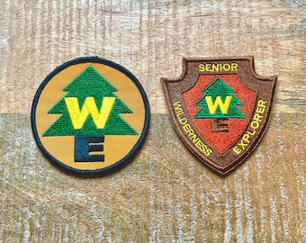 May include: Two embroidered patches featuring a green pine tree with a yellow "W" and "E" inside. The left patch is round with an orange background and black trim. The right patch is a brown shield shape with a red background and the text "Senior Wilderness Explorer".